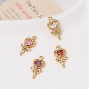 May include: Four gold-colored charms with heart-shaped designs. Each charm features a heart-shaped gemstone in pink, clear, red, and purple, accented with a bow and crystal details. The charms are on a white surface.
