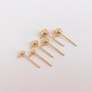 May include: Six gold-colored stud earrings with a ball-shaped top and a straight post. The earrings are arranged diagonally on a white surface, with varying sizes of the ball-shaped top.