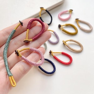 May include: A collection of colorful hair ties in various shades, including pink, red, and blue. Each tie features a looped design with a gold-colored accent and a small, dark-colored button. The ties are arranged on a light background.