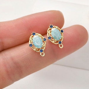 Può includere: Due ciondoli per orecchini ovali dorati con una pietra blu chiaro circondata da un design a filigrana decorativo e piccoli accenti blu.