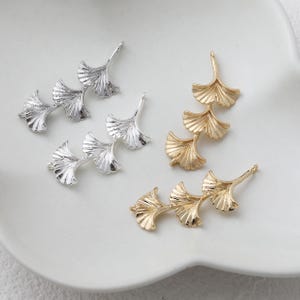 May include: Close-up of several metal ginkgo leaf charms in silver and gold. Each charm features three stylized leaves connected in a row, with a small loop at the top for attaching to jewelry. The charms are displayed on a white surface.