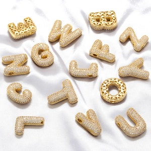 Może przedstawiać: Zestaw złotych zawieszek z literami alfabetu, każda litera jest ozdobiona przezroczystymi cyrkoniami. Zawieszki idealnie nadają się do dodania spersonalizowanego akcentu do projektów biżuterii.