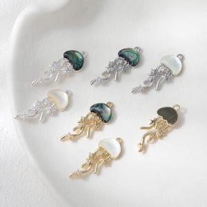 May include: A collection of jellyfish charms in silver and gold tones. The charms feature iridescent, white, and gold-colored tops with delicate, flowing tentacles. Some charms are adorned with small, sparkling accents.