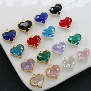 Puede incluir: Una colección de dijes en forma de corazón en varios colores, incluyendo azul, rojo, verde y morado. Cada dije tiene un borde dorado y un pequeño lazo para sujetar a la joyería. Los dijes se muestran sobre una superficie blanca.