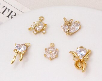 May include: Five gold-colored charms with clear, sparkling crystal accents. The charms include a heart with a bow, a heart with a butterfly, a heart with a ribbon, a geometric shape, and a charm with a floral design.