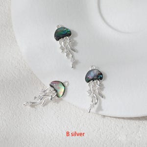 May include: Three silver-toned jellyfish charms with iridescent abalone shell caps and flowing tentacles. The charms have a loop at the top for attaching to jewelry. The text "B silver" is at the bottom.
