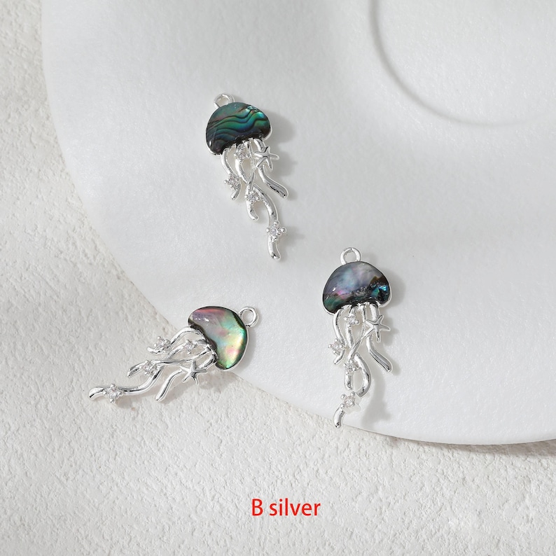 May include: Three silver-toned jellyfish charms with iridescent abalone shell caps and flowing tentacles. The charms have a loop at the top for attaching to jewelry. The text "B silver" is at the bottom.