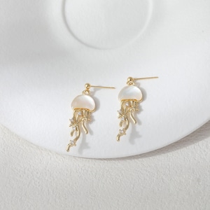May include: A pair of gold-colored jellyfish earrings. Each earring features a white, shell-like top and dangling tendrils adorned with small, clear crystals. The earrings are set against a white background.