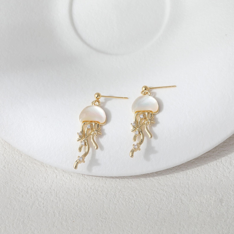 May include: A pair of gold-colored jellyfish earrings. Each earring features a white, shell-like top and dangling tendrils adorned with small, clear crystals. The earrings are set against a white background.