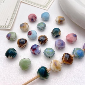 May include: Assortment of colourful ceramic shell-shaped beads with various floral patterns. The beads are arranged on a white surface and some are strung on a wooden skewer.