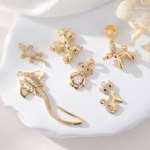 May include: An assortment of gold-toned charms, featuring a lizard, a teddy bear, a balloon dog, a cat, a stingray, and a cross. Each charm is adorned with tiny, clear crystals, enhancing the detail of these jewellery components.