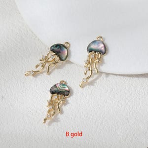 May include: Three gold-colored jellyfish charms with iridescent, shell-like caps and dangling tendrils. Each charm features small, clear crystals and a loop for attaching to jewelry. The text "B gold" is visible at the bottom.