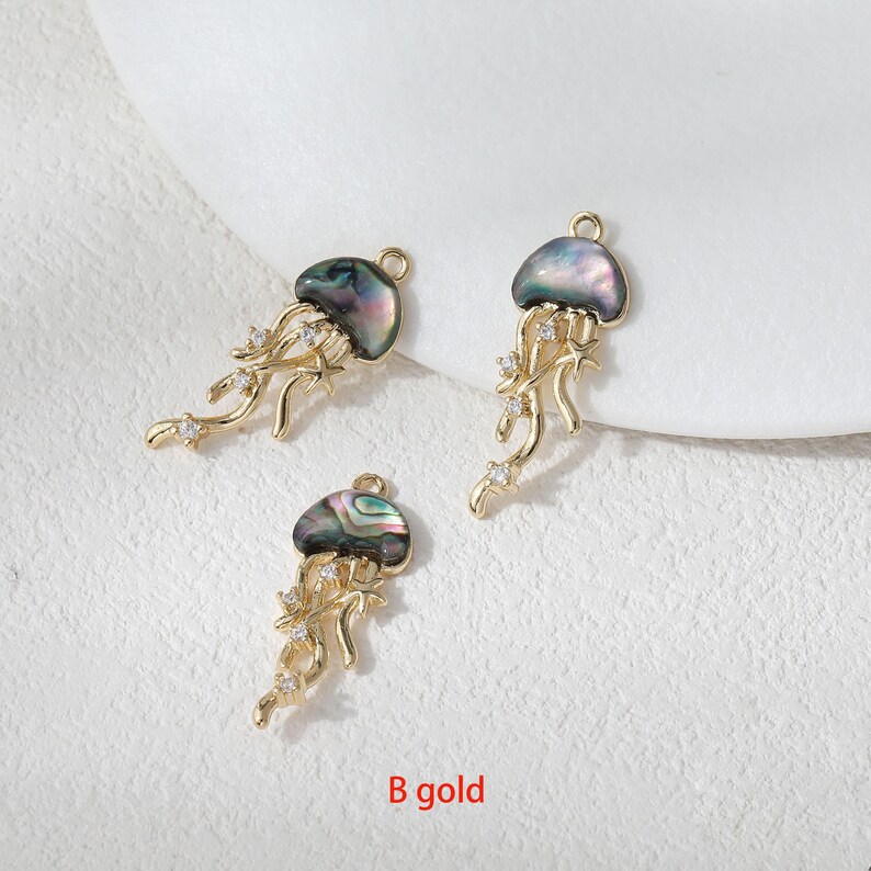 May include: Three gold-colored jellyfish charms with iridescent, shell-like caps and dangling tendrils. Each charm features small, clear crystals and a loop for attaching to jewelry. The text "B gold" is visible at the bottom.
