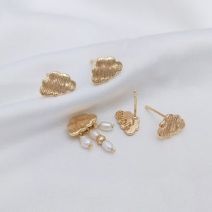 May include: Three pairs of gold cloud earrings. One pair features a cloud with a dangling pearl and bead charm. Another pair features a cloud with the word "cloud" inscribed on it.