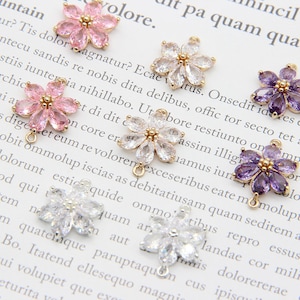 May include: Assortment of flower-shaped charms with gold-tone jump rings. The charms are made of clear, pink, and purple rhinestones.