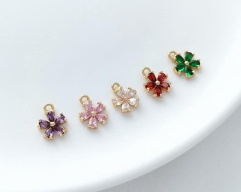 May include: Five gold flower charms with different colored rhinestones. The charms are arranged in a row on a white surface. The colors of the rhinestones are purple, pink, white, red, and green.