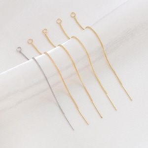 Può includere: Una collezione di orecchini a catena delicati in tonalità oro e argento. Ogni orecchino presenta una catena sottile con un piccolo anello in alto e una barra dritta in basso. Presentati su una superficie bianca.