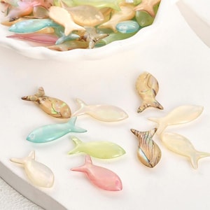 Può includere: Una collezione di perline a forma di pesce, iridescenti e colorate, in varie tonalità di rosa, verde, blu e beige. Alcune perline sono sparse su una superficie bianca, altre in una ciotola bianca. Finitura lucida.