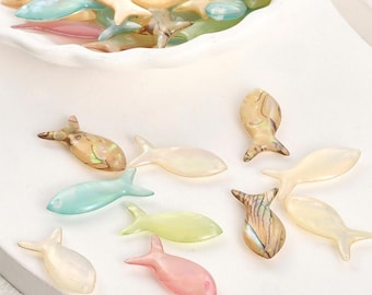 May include: A collection of colorful, iridescent fish-shaped beads in various shades of pink, green, blue, and beige. Some beads are scattered on a white surface, while others are in a white bowl. The beads have a glossy finish.
