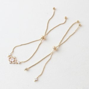 Pode incluir: Um bracelete ajustável de ouro com um design delicado de cristal transparente. O bracelete apresenta um pequeno cristal quadrado no centro, rodeado por cristais menores.