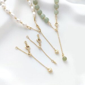 Peut inclure: Quatre chaînes en or de différentes longueurs, chacune ornée de petites boules dorées et se terminant par un pendentif en forme de cœur ou une perle de jade verte.