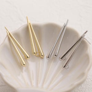 May include: Three pairs of earring findings, two gold and one silver, in the shape of a teardrop. The findings are on a white surface.