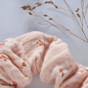 Könnte beinhalten: Nahaufnahme eines hellrosa Haargummis mit kleinen gestickten roten und grünen floralen Details. Das Haargummi ist aus einem weichen, strukturierten Stoff gefertigt. Getrocknetes Gras ist im Hintergrund.