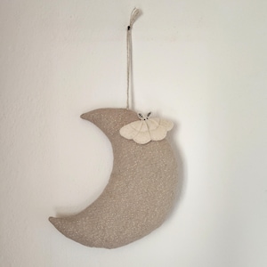 Könnte beinhalten: Ein halbmondförmiger Wandschmuck aus beigefarbenem Stoff mit glitzernder Textur. Auf der Oberfläche ist ein cremefarbener Schmetterling angebracht. Der Mond wird von einer dünnen, naturfarbenen Schnur gehalten.