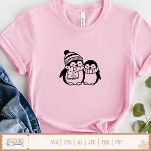 Puede incluir: Una camiseta de manga corta de color rosa claro con un gr&aacute;fico negro de dos ping&uuml;inos de pie uno al lado del otro. El ping&uuml;ino m&aacute;s grande lleva un gorro a rayas y una bufanda con corazones, mientras que el m&aacute;s peque&ntilde;o lleva una bufanda simple.