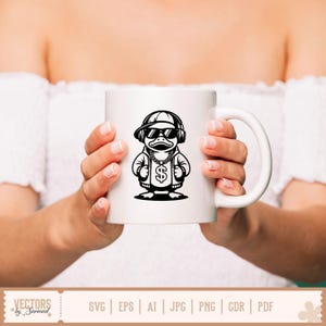 K&ouml;nnte beinhalten: Wei&szlig;e Keramiktasse mit einer Schwarz-Wei&szlig;-Illustration einer Ente mit Sonnenbrille, Kappe und Kopfh&ouml;rern. Die Ente hat eine goldene Kette mit einem Dollarzeichen-Anh&auml;nger. Die Tasse wird von einer Person gehalten, die ein wei&szlig;es Oberteil tr&auml;gt.