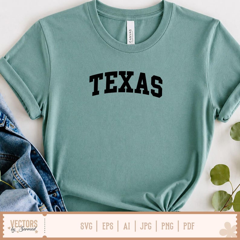 May include: A light teal t-shirt with the word "TEXAS" printed in bold, black letters across the chest. The shirt is made of a soft material and has a classic crew neck. A denim jacket is partially visible on the left side of the shirt.