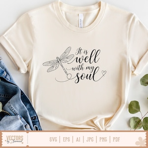 Puede incluir: Camiseta color crema con una ilustraci&oacute;n de lib&eacute;lula negra y la frase "It is well with my soul" en fuente cursiva. El dise&ntilde;o incluye un peque&ntilde;o coraz&oacute;n. La camiseta est&aacute; hecha de un material suave.