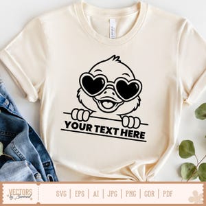 K&ouml;nnte beinhalten: Cremefarbenes T-Shirt mit einer schwarzen Cartoon-Ente, die eine herzf&ouml;rmige Sonnenbrille tr&auml;gt. Die Ente h&auml;lt ein Banner mit dem Text "YOUR TEXT HERE". Das Shirt hat kurze &Auml;rmel und einen Rundhalsausschnitt.