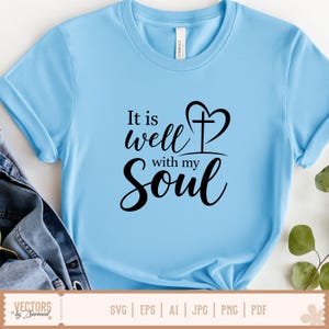 Puede incluir: Camiseta azul claro con la frase "It is well with my Soul" en escritura negra. El dise&ntilde;o incluye una cruz en forma de coraz&oacute;n. La camiseta es de material suave y tiene mangas cortas.