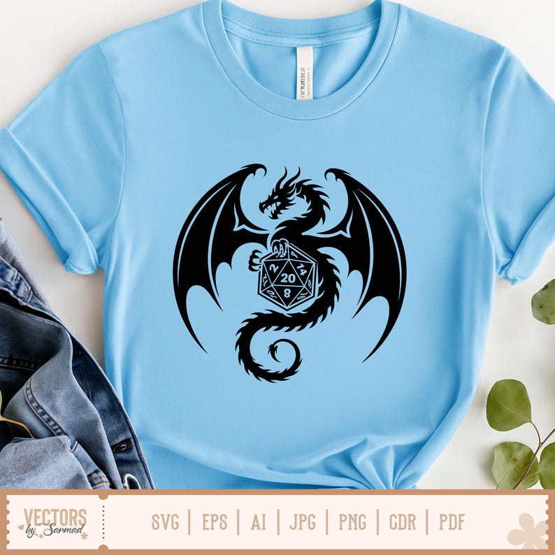 May include: Pale blue t-shirt with a black dragon graphic holding a twenty-sided die. The dragon has large wings and a detailed head. The die shows the numbers 20 and 8. The shirt is made of a soft material.