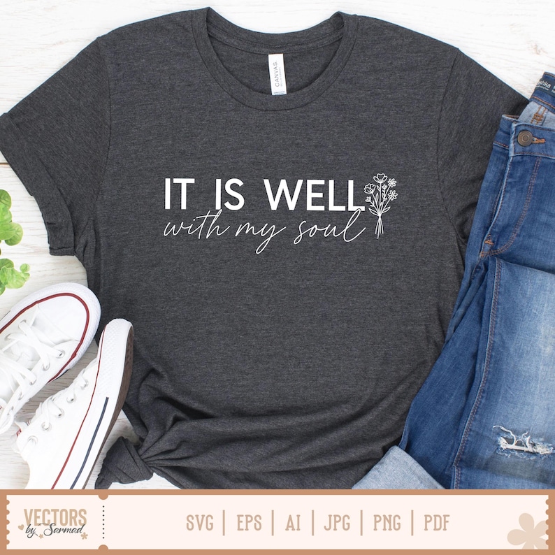Puede incluir: Camiseta gris oscuro con el texto blanco "IT IS WELL with my soul". Un peque&ntilde;o dise&ntilde;o floral est&aacute; a la derecha del texto. Zapatillas blancas y vaqueros azules son visibles.