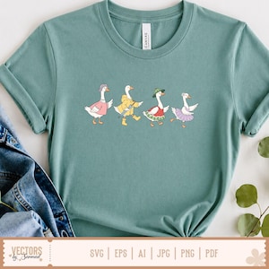 May include: A dusty teal crew neck t-shirt featuring a graphic of four white geese walking in a line. The geese are dressed in various outfits including a pink jumper, a yellow raincoat with an umbrella, a watermelon-patterned dress, and a purple tutu.