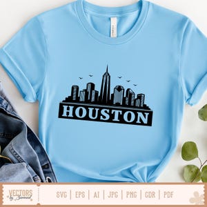 Peut inclure: T-shirt bleu clair avec une silhouette noire de l'horizon de Houston et le nom de la ville en lettres grasses. Le design comprend des oiseaux. Le t-shirt a des manches courtes et un col rond.
