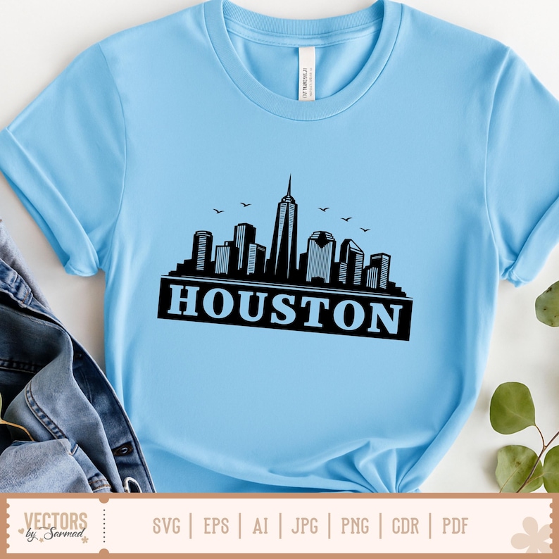 Peut inclure: T-shirt bleu clair avec une silhouette noire de l'horizon de Houston et le nom de la ville en lettres grasses. Le design comprend des oiseaux. Le t-shirt a des manches courtes et un col rond.