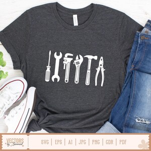 May include: Dark gray t-shirt featuring a white graphic of various tools, including a screwdriver, wrench, adjustable wrench, hammer, and pliers. The shirt is paired with blue jeans and white sneakers. The word "CANVAS" is printed on the shirt.