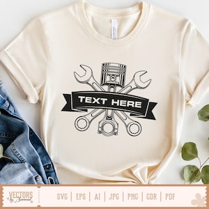 May include: Cream-colored t-shirt featuring a black graphic of crossed wrenches, a piston, and a connecting rod, with a banner reading "TEXT HERE." The shirt has short sleeves and a crew neck. The bottom of the shirt has a tag with the text "VECTORS by Sarmad."