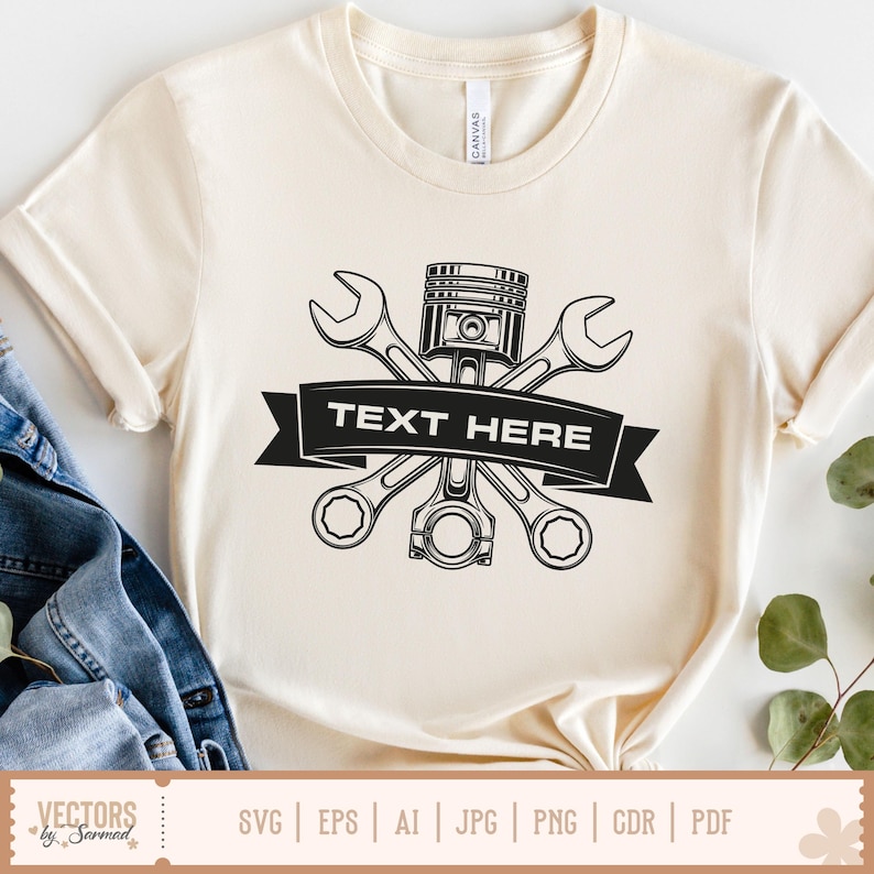 May include: Cream-colored t-shirt featuring a black graphic of crossed wrenches, a piston, and a connecting rod, with a banner reading "TEXT HERE." The shirt has short sleeves and a crew neck. The bottom of the shirt has a tag with the text "VECTORS by Sarmad."