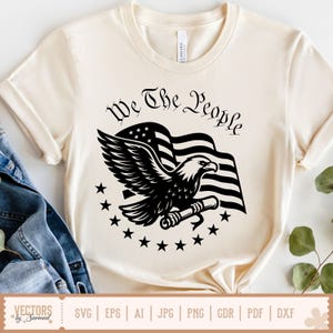 以下が含まれることがあります： 白いTシャツに、黒いデザインが施されています。デザインには、巻物を持った鷲が描かれており、巻物には「We The People」という文字が書かれています。背景にはアメリカの国旗が描かれており、デザイン全体を星が囲んでいます。