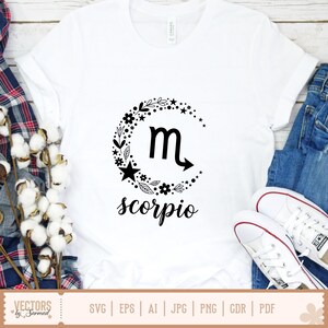 Op de afbeelding: Wit T-shirt met een zwart Schorpioen dierenriem ontwerp. Het ontwerp bevat het Schorpioen symbool, bloemen en ster accenten, en het woord "scorpio" in script. Het shirt wordt gecombineerd met een blauwe jeans en witte sneakers.