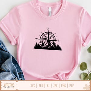 以下が含まれることがあります： 胸元に黒いグラフィックプリントが施されたライトピンクの半袖Tシャツ。デザインは、山頂のシルエットと密集した松林の上に配置されたコンパスローズで、N、E、W、NW、NEの文字が含まれています。
