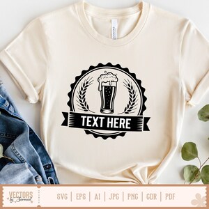 May include: Cream-colored t-shirt featuring a black beer glass graphic with foam, surrounded by wheat stalks and a banner that reads "TEXT HERE." The shirt is made of soft material and has rolled-up sleeves.