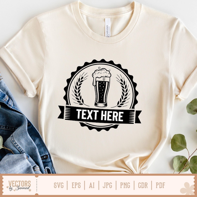May include: Cream-colored t-shirt featuring a black beer glass graphic with foam, surrounded by wheat stalks and a banner that reads "TEXT HERE." The shirt is made of soft material and has rolled-up sleeves.