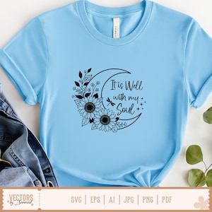 Puede incluir: Camiseta azul claro con un dise&ntilde;o gr&aacute;fico negro. El dise&ntilde;o presenta una luna creciente, girasoles y el texto "It is Well with my Soul". La camiseta es de material suave.