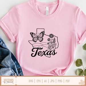 May include: Pink t-shirt with a black graphic of the state of Texas, a butterfly, and floral elements. The word "Texas" is written in a cursive font below the graphic. The shirt is made of a soft material.