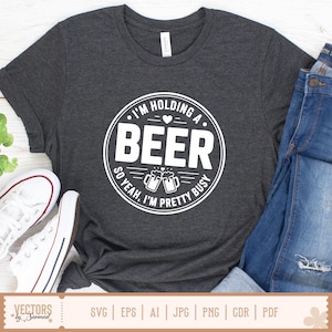 May include: Dark gray t-shirt with a white circular graphic that reads "I'M HOLDING A BEER SO YEAH, I'M PRETTY BUSY." The design includes two beer mugs. The shirt is paired with blue jeans and white sneakers.